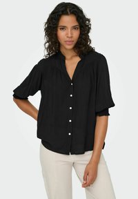 Sort bluse med korte, flagrende ærmer, knaplukning foran og blødt, let materiale. Udvikler sig med sin afslappede pasform og bløde folder ved skuldrene.