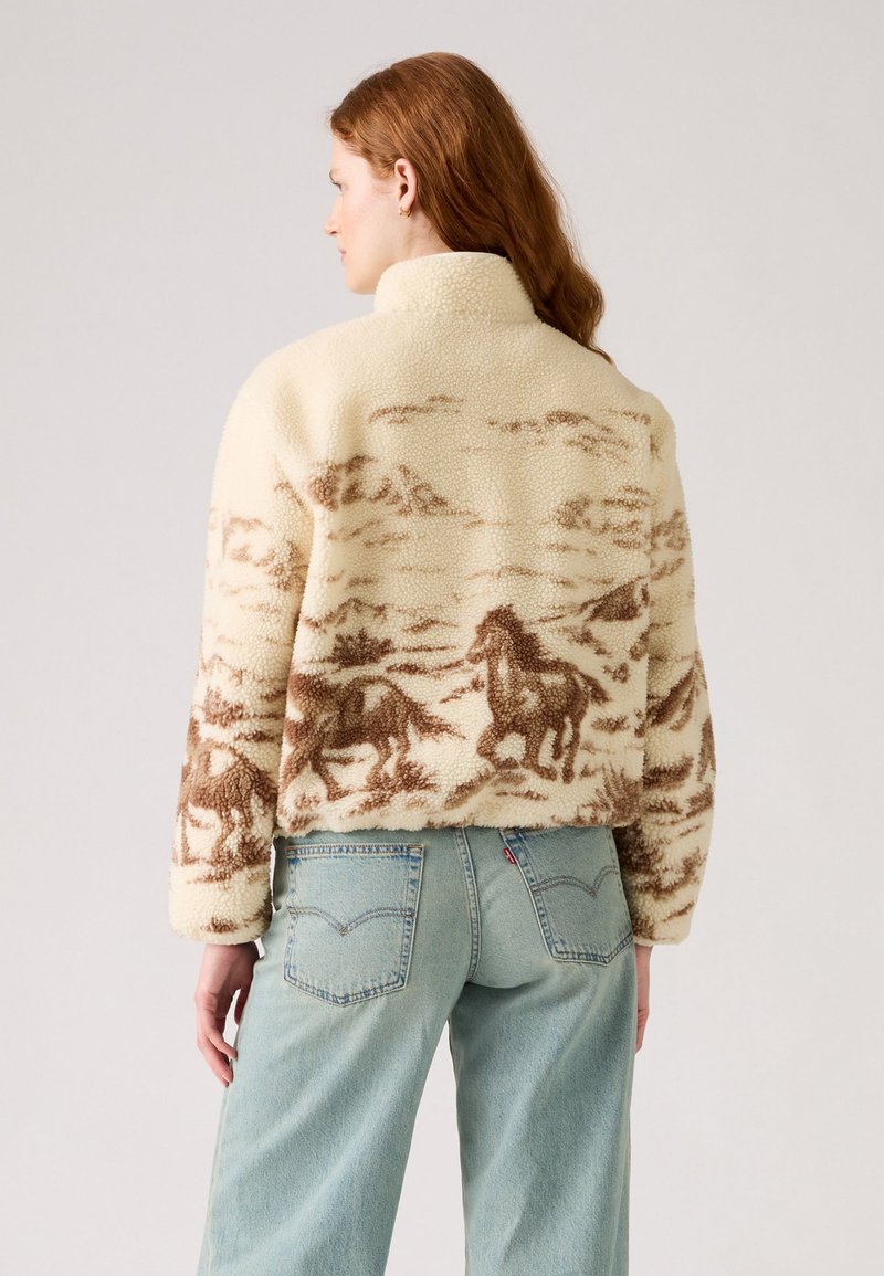 Cremfarbener Fleece-Jacke mit hohem Kragen, mit braunem Pferdemuster. Lockere Passform, gerippte Bündchen und Saum. Ein Paar hellblaue Jeans.