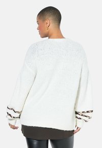 Bílý pletený svetr s širokými rukávy, s leopardím potiskem na manžetách. Měkká textura a volný střih.