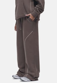 Bruine oversized hoodie met een kangoeroe pocket en witte stiksel, gecombineerd met bijpassende losse sweatpants en grijze sportschoenen.