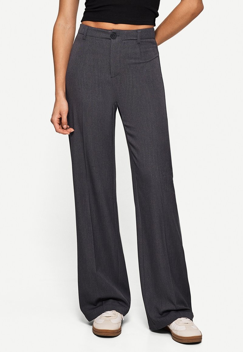 Personne portant un pantalon large gris foncé taille haute avec fermeture à boutons, associé à des baskets blanches et un top court noir.