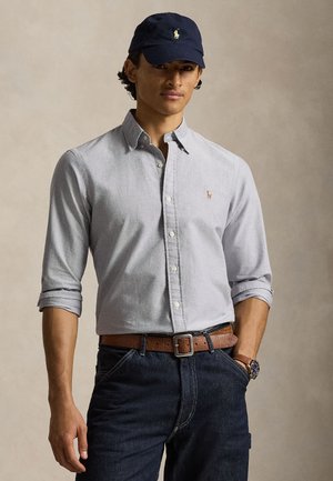 Νέος άνδρας που φοράει ανοιχτό γκρι πουκάμισο με κουμπιά, μπλε σκουρόχρωμο καπέλο μπέιζμπολ, σκούρα τζιν με καφέ ζώνη και ρολόι χειρός, στέκεται μπροστά σε απλό φόντο.