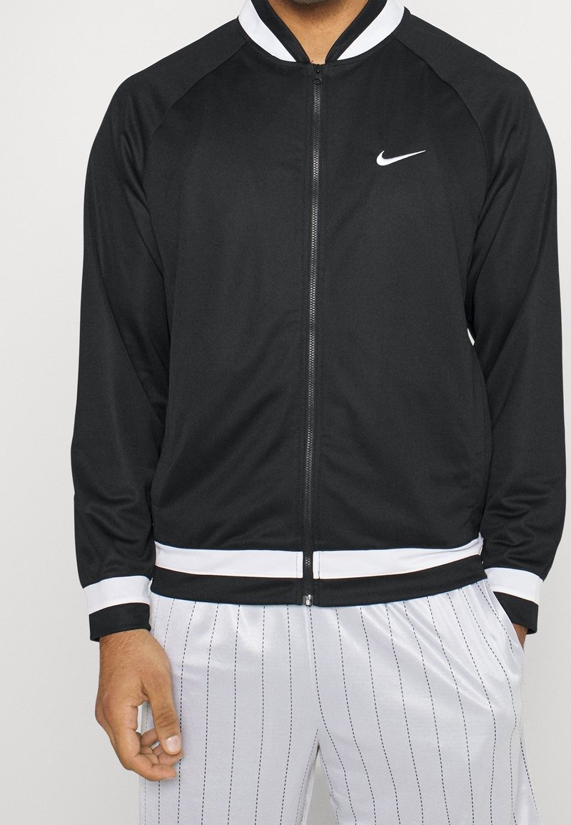 Veste noire zippée avec des accents blancs, dotée d'un col montant et de poignets élastiques, fabriquée en matériau lisse et léger.