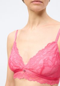 Γυναίκα που φοράει ροζ δαντελένιο μπραλέτο με λουλουδάτα σχέδια και λεπτές ρυθμιζόμενες τιράντες, σε απλό λευκό φόντο.