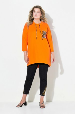 Tunique orange avec une poche à imprimé léopard, associée à un pantalon noir avec des accents dorés. Manches trois-quarts et design à ourlet arrondi.