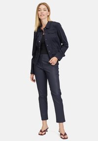 Marineblaue Jeansjacke mit taillierter Passform, Fronttaschen und silbernen Beschlägen, kombiniert mit schmal geschnittenen marineblauen Hose und kastanienbraunen Sandalen.