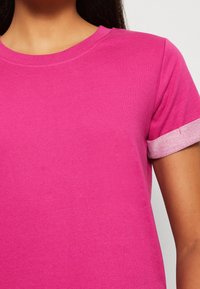 Różowa koszulka z krótkim rękawem, wykonana z miękkiego materiału, z prążkowanym dekoltem i kontrastującymi, teksturowanymi mankietami w jaśniejszym odcieniu.