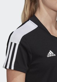 Svart Adidas sporttröja med vit axelpanel och tre vita ränder på ärmen, bärs av person med blont hår i hästsvans.