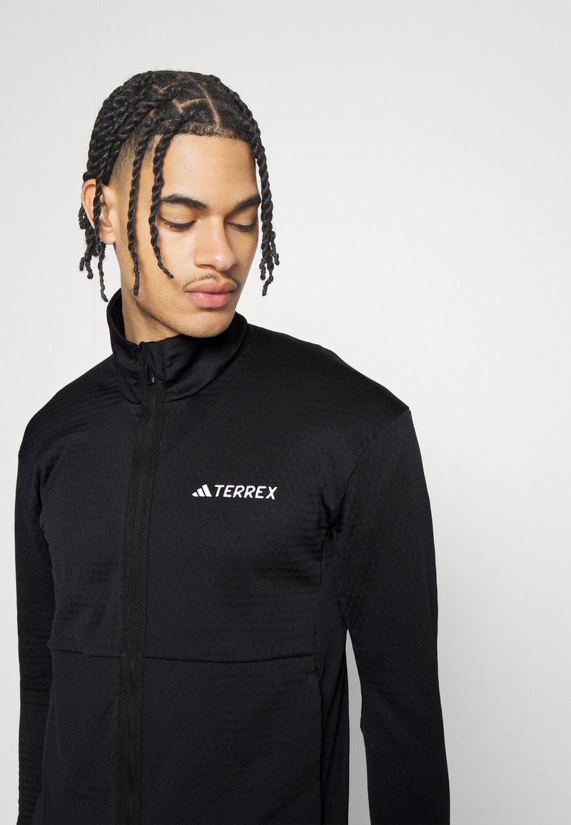 Jonge man met gevlochten haar in een zwarte Adidas Terrex jack met ritssluiting die omlaag kijkt tegen een effen lichte achtergrond.