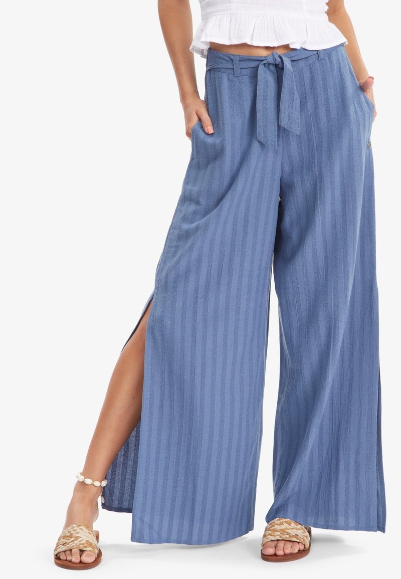 Pantalon bleu clair à jambes larges avec rayures verticales, ceinture nouée, fentes latérales et poches. Confectionné dans un tissu doux et texturé.