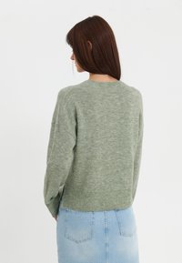 Pull en laine vert avec un ourlet et des poignets côtelés, présentant une texture douce et une coupe décontractée, associé à une jupe en denim bleu.
