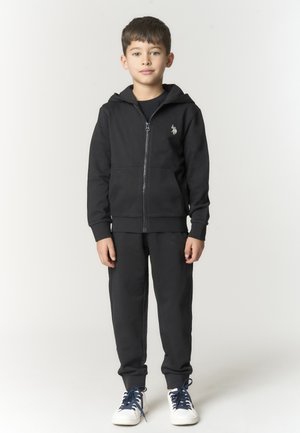 Ragazzo in piedi che indossa una felpa nera con zip, pantaloni neri e scarpe da ginnastica bianche con lacci blu scuro su uno sfondo chiaro uniforme.