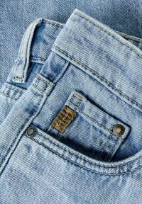 Primo piano di jeans in denim azzurro chiaro che mostrano la tasca anteriore con rivetti metallici e un piccolo etichetta in tessuto marrone cucita all'interno della tasca.