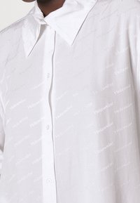 Dettaglio ravvicinato di una camicia bianca con colletto ampio e un sottile motivo ripetuto con il testo "Holzweiler" sul tessuto.