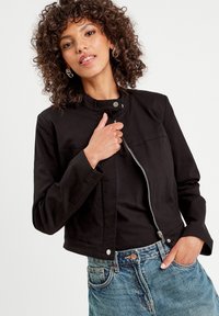 Black cropped jacket with a high collar, silver zipper, and snap closure; paired with denim shorts featuring a classic blue wash and front pockets.
