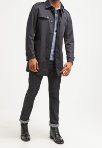 Trench-coat bleu marine foncé avec des boutons et des poignets ajustables, associé à une chemise bleu clair, un jean noir et des bottes à lacets noires.