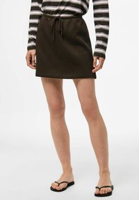 Mini gonna marrone con cordino, realizzata in tessuto morbido, caratterizzata da una consistenza liscia e un design semplice, abbinata a infradito nere.