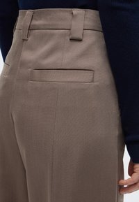 Κοντινό πλάνο ενός ατόμου που φοράει ταφτά παντελόνια με ζώνη και πίσω τσέπη με φάσα, σε συνδυασμό με σκουρόχρωμο μακρυμάνικο τοπ.