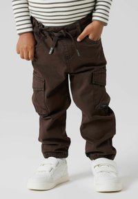 Brune cargobukser med elastiske mansjetter, sidelommer og snor i midjen, kombinert med hvite velcro-sneakers. Stripete topp delvis synlig.