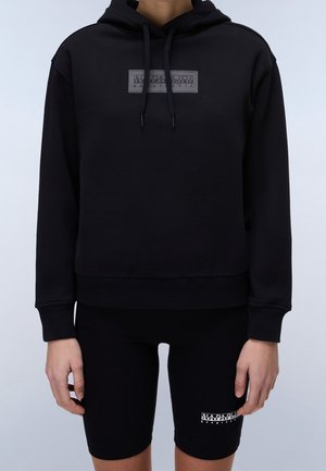 Personne portant un sweat à capuche noir avec un logo sur le devant et un cuissard de cyclisme assorti noir avec un logo sur la cuisse gauche, se tenant devant un fond uni.