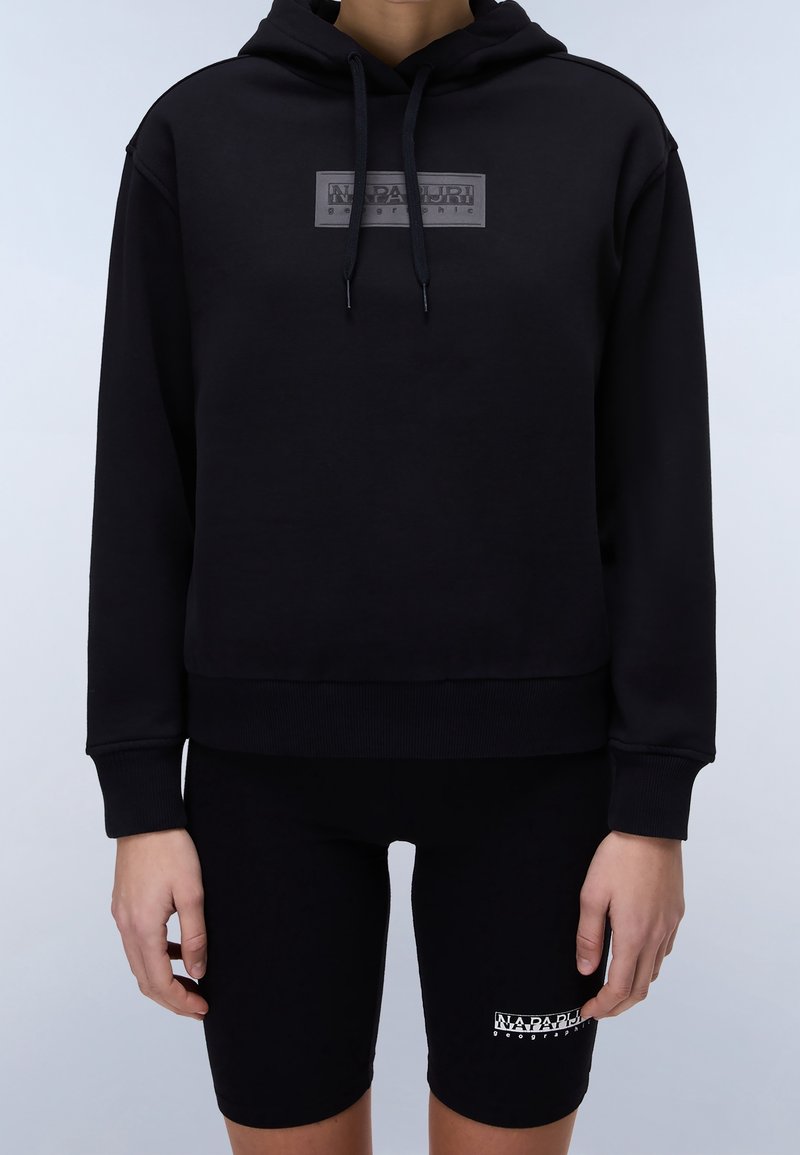 Personne portant un sweat à capuche noir avec un logo sur le devant et un cuissard de cyclisme assorti noir avec un logo sur la cuisse gauche, se tenant devant un fond uni.