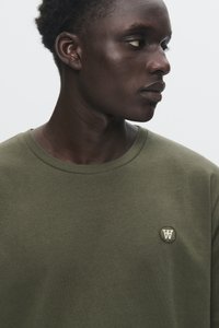 Young Black man wearing an olive green shirt with a small circular emblem on the chest, looking to his right against a plain background.