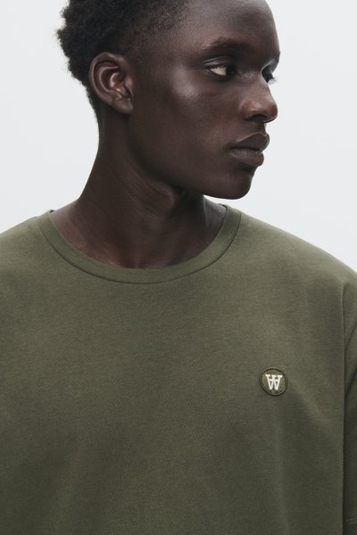 Young Black man wearing an olive green shirt with a small circular emblem on the chest, looking to his right against a plain background.