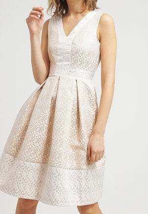 Mouwloze beige jurk met V-hals, subtiel polkadotpatroon, getailleerde taille en uitlopende geplooide rok, gedragen door een gedeeltelijk zichtbare persoon.