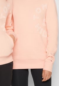Felpa rosa chiaro realizzata in tessuto morbido, con testo bianco e una tasca frontale. I polsini hanno un piccolo logo come dettaglio.