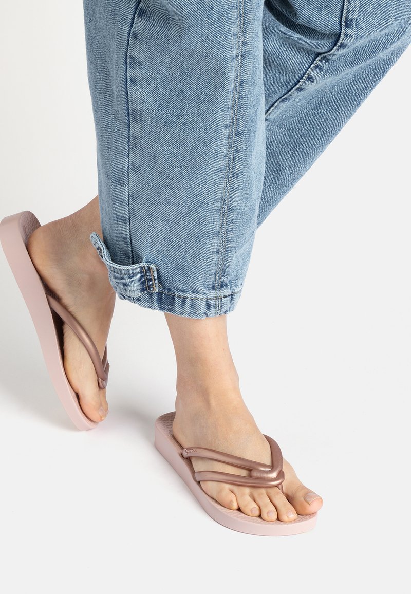 Rosa Flip-Flops mit einer leichten Sohle und einem strukturierten Fußbett, ausgestattet mit zwei geschwungenen Riemen in einem kontrastierenden metallischen Farbton. Jeans sind oben sichtbar.