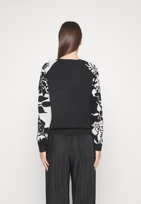 Svart tröja med förlängda ärmar som har kontrasterande svarta och vita blommönster. Ribbad mudd och nederkant; slät textur.