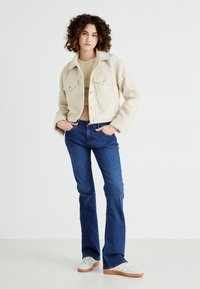 Cream, cropped fleece jacket with front pockets and silver buttons, paired with blue flared jeans and white sneakers with brown soles.