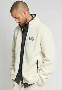 Fleecejacke in Cremefarbe mit marineblauem Besatz. Mit einem Reißverschluss vorne, zwei seitlichen Taschen und einem Brustlogo-Patch. Weiche Textur.