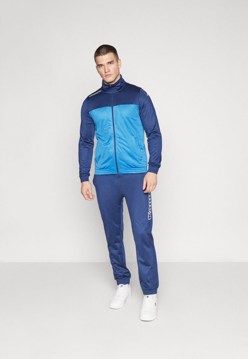 Tuta sportiva blu e navy con giacca zip-up e pantaloni elastici, texture liscia, con dettagli del logo su giacca e pantaloni.