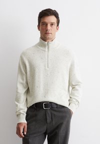 Helles beige Pullover mit hohem Kragen, Reißverschluss und gestrickter Textur, kombiniert mit grauen Hosen und einem schwarzen Gürtel.
