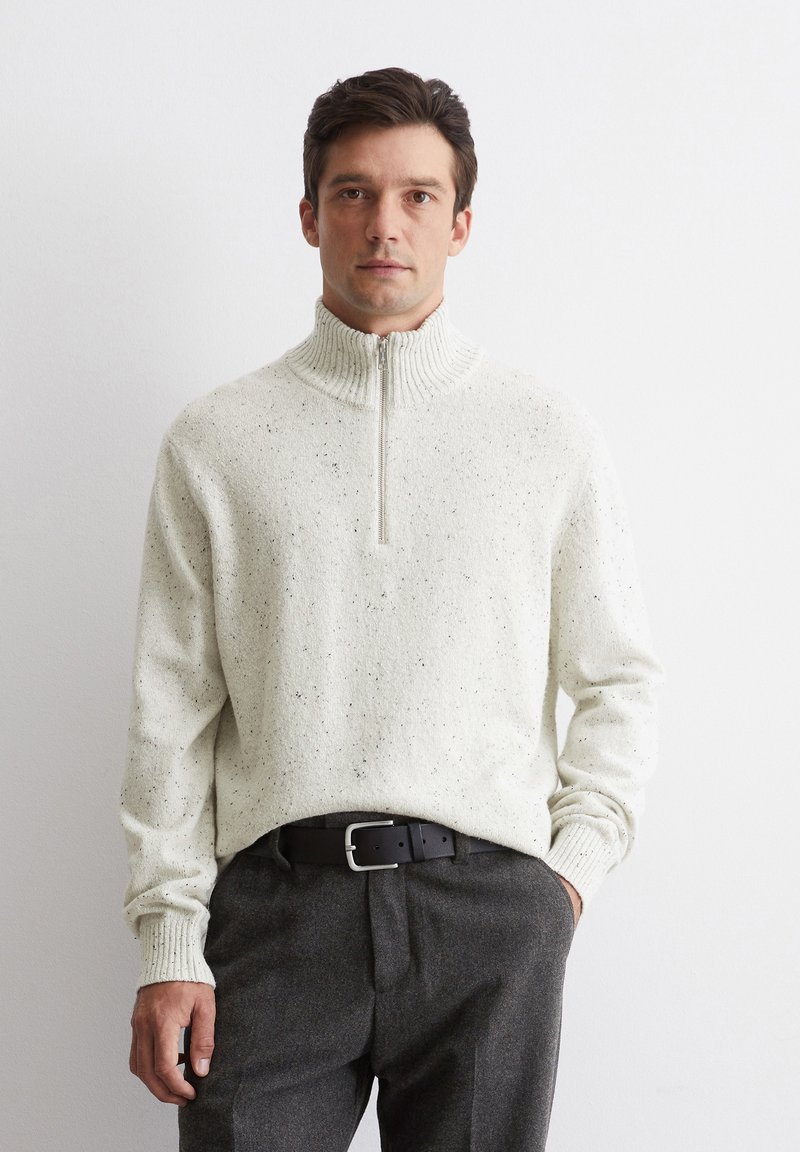 Helles beige Pullover mit hohem Kragen, Reißverschluss und gestrickter Textur, kombiniert mit grauen Hosen und einem schwarzen Gürtel.