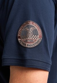 Marinblå t-shirt med kort ärm och ett runt, broderat märke med en örn, rutmönster och textdetaljer om Daytona.