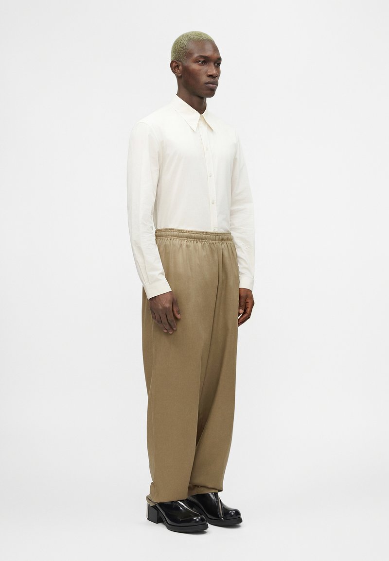 Homme aux cheveux blonds courts portant une chemise blanche boutonnée, un pantalon beige ample et des chaussures noires brillantes, debout devant un fond uni.