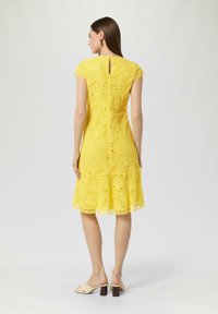 Gelbes Spitzenkleid mit tailliertem Oberteil und ausgestellt Rock mit gewelltem Saum. Enthält ein Schlüsselaugen-Detail im Rücken und kurze Ärmel.