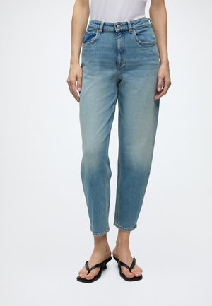 Une femme portant un jean taille haute bleu clair et des sandales noires à talons ouverts, debout devant un fond uni.