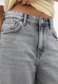 Jean gris clair taille haute avec une finition délavée, cinq poches et une fermeture à bouton métallique. Détail d'usure subtil près de la poche inférieure gauche.