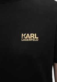 Μαύρο μπλουζάκι με χρυσό λογότυπο «KARL LAGERFELD», που διαθέτει τη σιλουέτα του Karl Lagerfeld ενσωματωμένη στο γράμμα Κ.