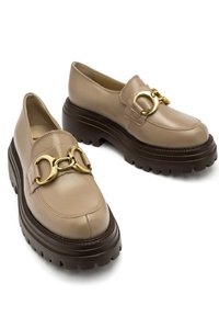 Beżowe skórzane mokasyny z masywną brązową podeszwą, z złotym metalowym zapięciem z przodu, gładkim wykończeniem i okrągłym noskiem.
