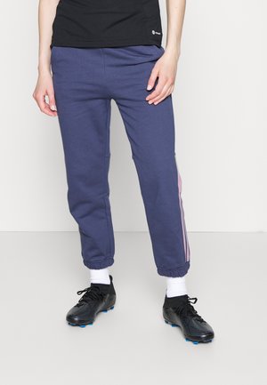 Person trägt marineblaue Jogginghose mit pinken Seitenstreifen, weiße Socken, schwarze Stollenschuhe und ein schwarzes Oberteil und steht vor einem einfarbigen Hintergrund.