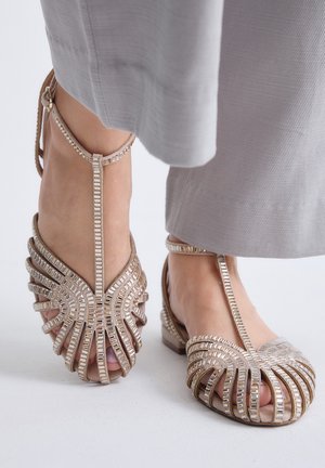 Pies con sandalias beige con tira en T y cuentas decorativas metálicas rectangulares combinadas con pantalones gris claro.