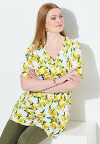 Tunic bianca con un vivace motivo di limoni gialli e foglie verdi. Maniche corte, scollo a V e vestibilità rilassata. Abbinata a pantaloni olive.