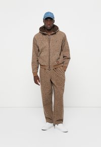 Bruine katoenen trainingspak met een luipaardprint, bestaande uit een hoodie met rits en een broek met trekkoord, gecombineerd met een lichtblauw pet en witte schoenen.