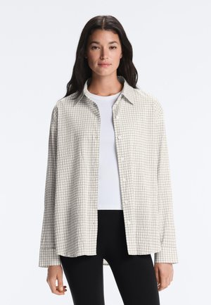 Žena s dlhými tmavými vlasmi má na sebe otvorenú svetlú károvanú košeľu cez biely top a čierne nohavice, stojí proti jednoliatemu pozadiu.