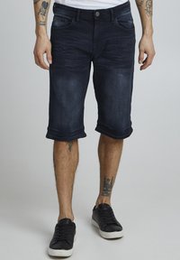 Shorts en denim foncé avec ourlets retroussés, design à cinq poches et fermeture à bouton. Portés avec des baskets noires sur un fond uni.