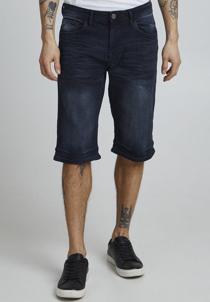 Shorts en denim foncé avec ourlets retroussés, design à cinq poches et fermeture à bouton. Portés avec des baskets noires sur un fond uni.